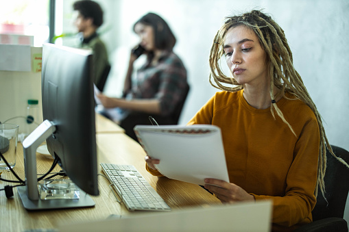 Young woman with dreadlocks working on plans in the office. There are people in the background.
