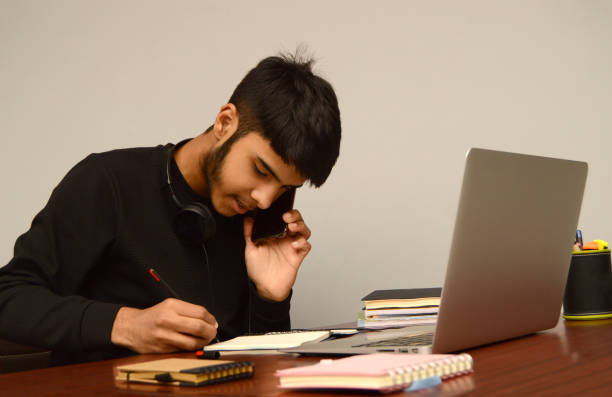 horizontal photograph of  one teenage boy or young male with black hair and beard  looking down at the notepad while writing and taking notes and talking over mobile phone, laptop screen busy multi tasking on his desk over grey background - homework teenager mobile phone school imagens e fotografias de stock
