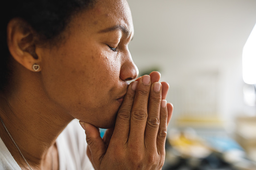 Profile view of distraught mid adult African American woman sitting with praying hands in front of face, eyes closed, battling anxiety and negative emotions cause of uncertainty of daily life.