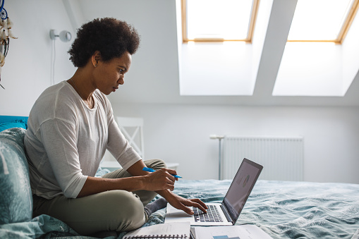 Profile view of diligent mid adult African American woman working remotely from her bedroom, using laptop while finishing a project.