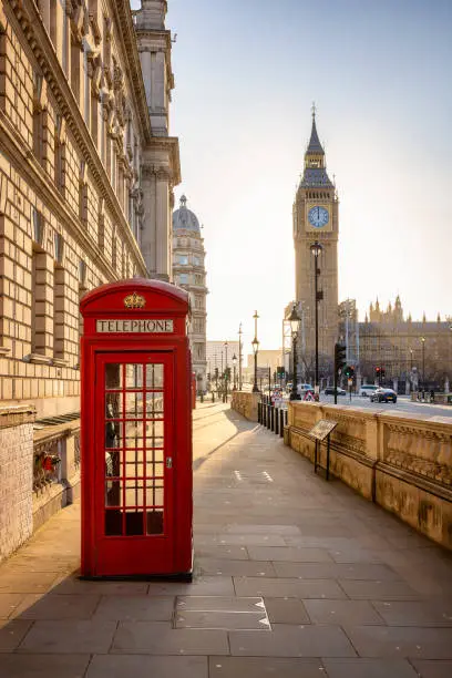 A classic, red telephone booth in front of the Big Ben clocktower in London, Westminster, during golden sunrise without people