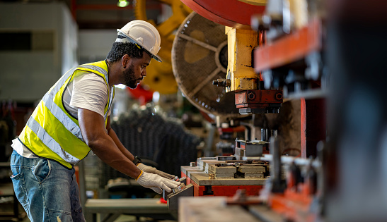 Trabajador industrial afroamericano está utilizando una máquina de prensa de potencia hidráulica para hacer piezas de metal y acero mientras trabaja dentro de la fábrica de techo galvanizado de chapa metálica para el concepto de la industria de seguri photo