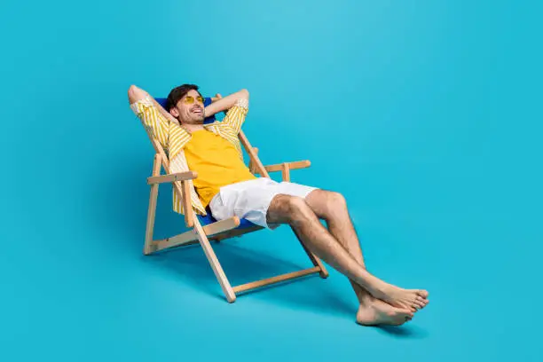 Full length profile side photo of positive guy, traveler relax rest beach sun bathing lie comfort deckchair stretch hands wear white shorts isolated over blue color background