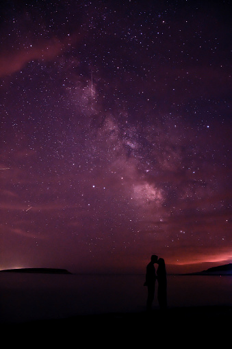 A couple ( a man and a woman) under the stars in the night looking at each other. Above the sea is the night sky painted in blue and purple with stars and milky way.