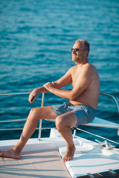 mężczyzna w okularach przeciwsłonecznych i kufrach siedzący na pokładzie i cieszący się światłem słonecznym na wakacjach żeglarskich - clear sky water sports and fitness yacht zdjęcia i obrazy z banku zdjęć