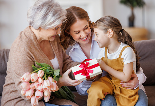 Happy International Women's Day. Smiling multi generational family daughter and granddaughter giving flowers  and gift to grandmother  cheerfully celebrate the spring holiday Mother's Day or grandparents day at home