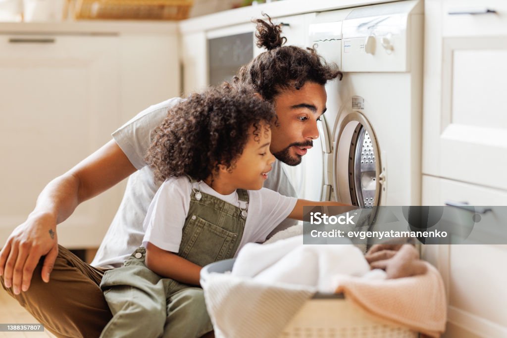 Son helping dad to load washing machine Cheerful black kid boy sitting on dad's lap and helping father at linen in basket while doing laundry near washing machine in flight kitchen in weekend at home Laundry Stock Photo