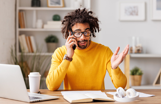 Friendly African American man in yellow sweatshirt and glasses raising arm and emotional speak while sitting at table and speaking on smartphone during work from home