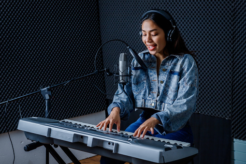 Happy cheerful pretty smiling of young Asian woman vocalist Wearing Headphones recording a song front of microphone and playing the keyboard during rehearsal of her band in a professional studio