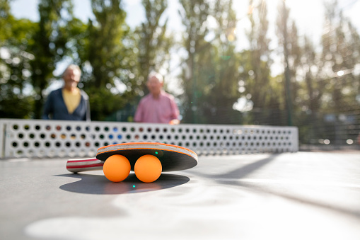 A racket leaning on two table tennis balls on a table tennis table in Newcastle Upon Tyne. There are two senior men on the opposite side of the table, out of focus, ready to play the game.