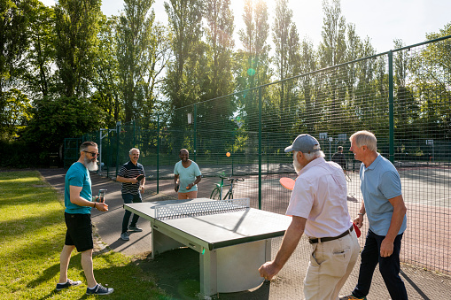 A group of senior men playing table tennis outdoors in a public park in Newcastle Upon Tyne, England. Four men are playing the game while another man watches them.