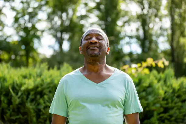 A senior man wearing a t-shirt, standing outdoors in nature in Newcastle upon Tyne, England. He is inhaling and exhaling in the fresh air.