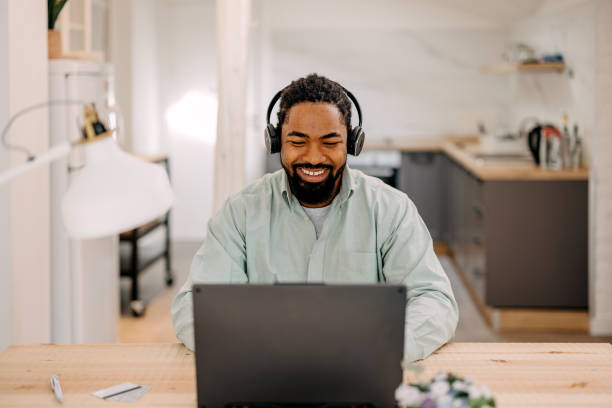 istock Smiling bearded African man using laptop at home while sitting the wooden table. 1388373123