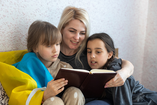 mother and two daughters are reading with the flag of ukraine in bed.