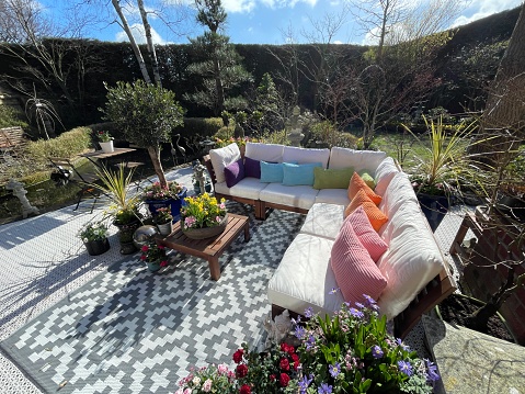 Stock photo showing ornamental Japanese-style garden with outdoor lounge area in Spring. Featuring a large expanse of white, interconnecting, white plastic decking tiles with outdoor patterned rug, providing a family space for outdoor hardwood, cushion covered seating.