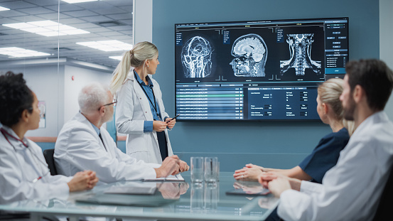 Hospital Conference Meeting Room: Female Neurologist Shows MRI Scan Brain Images on TV Screen, Team of Neuroscientists, Doctors Discuss Patient Treatment, Drug Research, Medicine Development