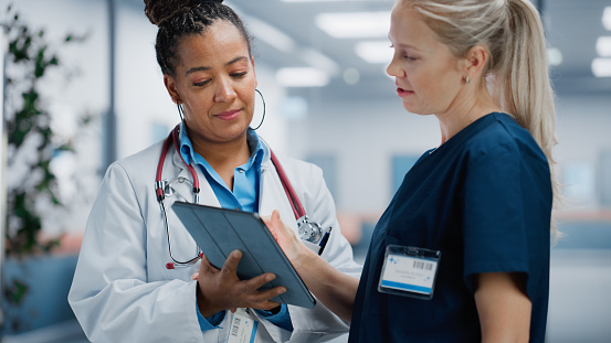 Medical Hospital: Professional Black Female Medical Doctor Talks with White Female Nurse Using Digital Tablet Computer. Two Health Care Specialists Discuss, Find Treatment Solution to Save Lives