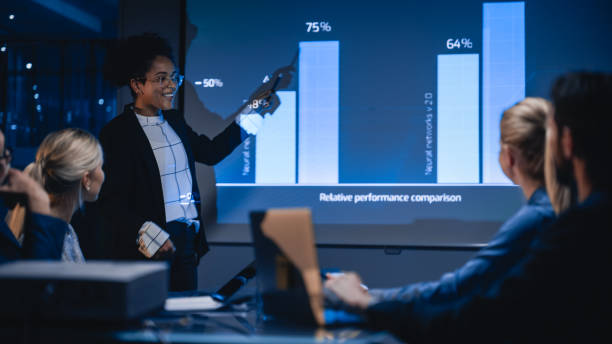istock e-Business Technology Conference Presentation: Innovative Black Businesswoman Talks about Revolutionary High-Tech Product. Projector Screen Shows Graphs, Infographics, AI, Big Data, Machine Learning 1388388097