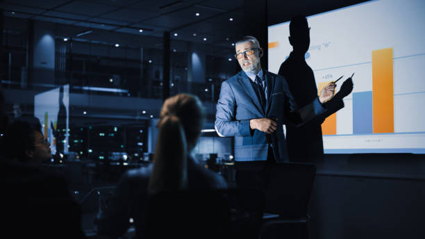 istock Conference Business Meeting Presentation: Chief Executive Officer Shows Data to Diverse Group of Investors, Businessspeople. Projector Screen Shows Graphs, Product Sales Growth, e-Commerce Analysis 1388388157