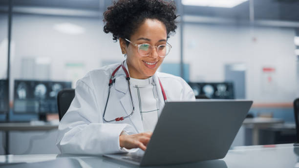 Hospital Doctors Office: Portrait of Young Promising Black Physician Works on Laptop Computer. Female Medical Professional in White Lab Coat Looking for Patient Treatment While Sitting at Her Desk Hospital Doctors Office: Portrait of Young Promising Black Physician Works on Laptop Computer. Female Medical Professional in White Lab Coat Looking for Patient Treatment While Sitting at Her Desk physician computer stock pictures, royalty-free photos & images