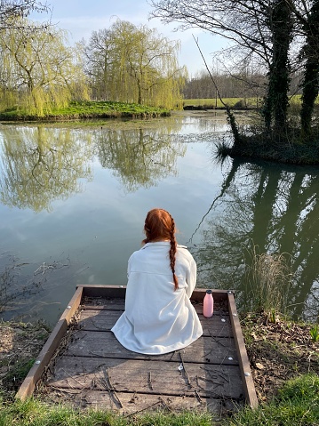 Stock photo showing the rear view of a young, redheaded woman wearing braids sitting on a bank of a lake in Springtime.