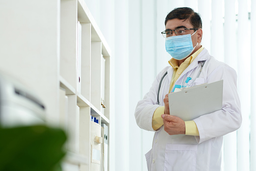 Doctor in medical mask searching for folder with medical history of patient on shelf