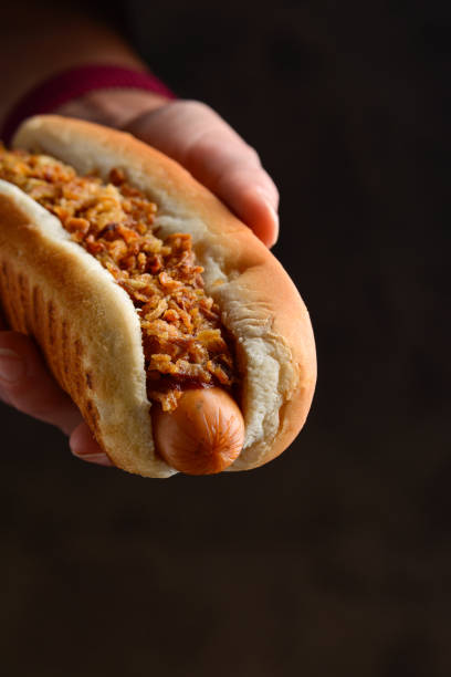 Hot dog with onion fries in a soft wheat bun in a woman's hand on a dark background. Sausage in the dough. Street food concept