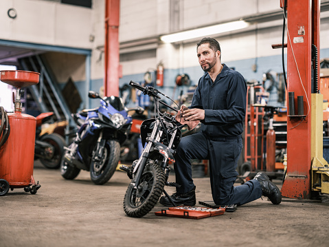 Mexican male mechanic in his shop. Mid adult man with beard and short hair, wearing work clothes. Interior of car and motorcycle repair shop during day.