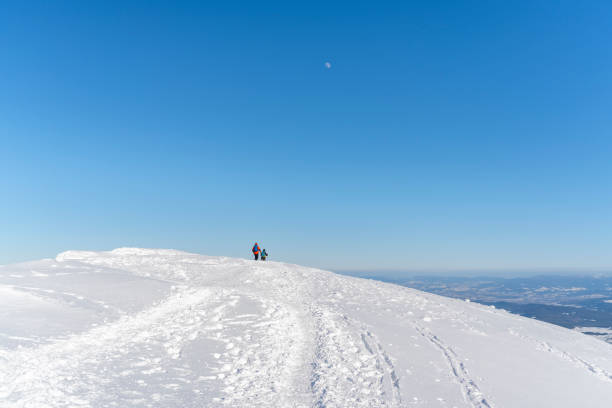 Couple of tourists on a sunny day on a snowy mountain trail, seen from a distance against the backdrop of a blue sky.