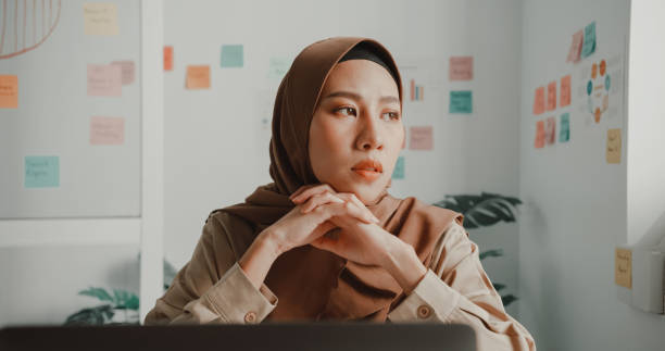 frustrierte junge asiatische muslimische geschäftsfrau im hijab, die an einem laptop am schreibtisch im büro arbeitet. - pensive business person looking up ideas stock-fotos und bilder