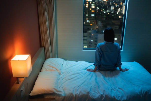 Rear view of woman sitting alone on bed in room and looking through window at night stock photo
