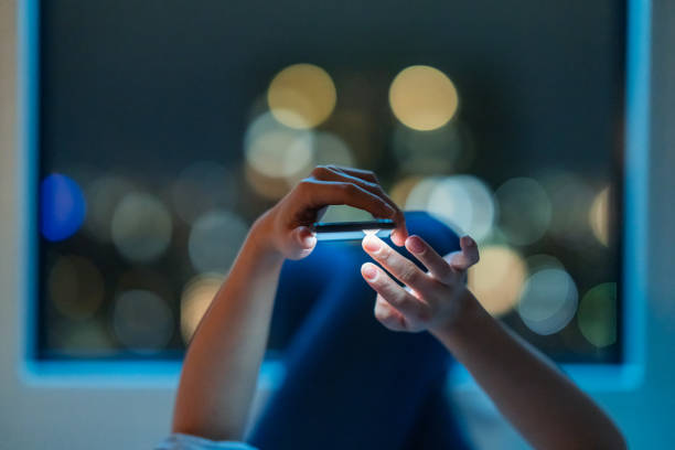 Woman lying down on bed and using smart phone at night A woman is lying down on a bed and using a smart phone at night. mobile phone lights up stock pictures, royalty-free photos & images