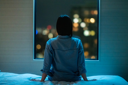 Vista trasera de la mujer sentada sola en la cama de la habitación y mirando a través de la ventana por la noche photo