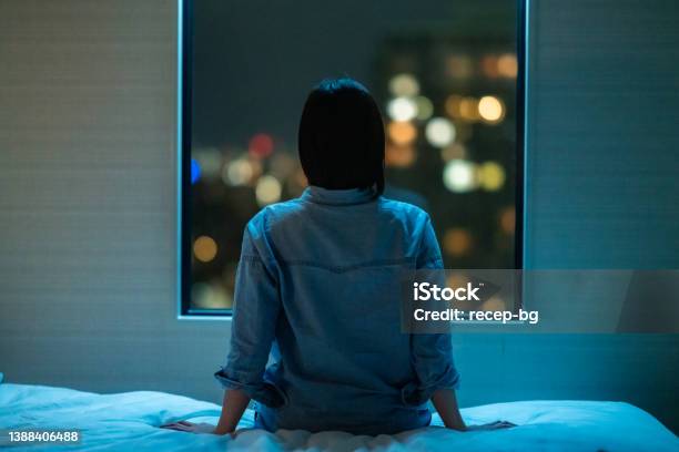 部屋のベッドに一人で座り、夜に窓から覗く女性の背面図