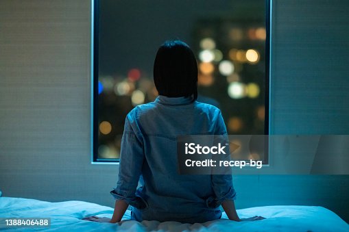 istock Rear view of woman sitting alone on bed in room and looking through window at night 1388406488