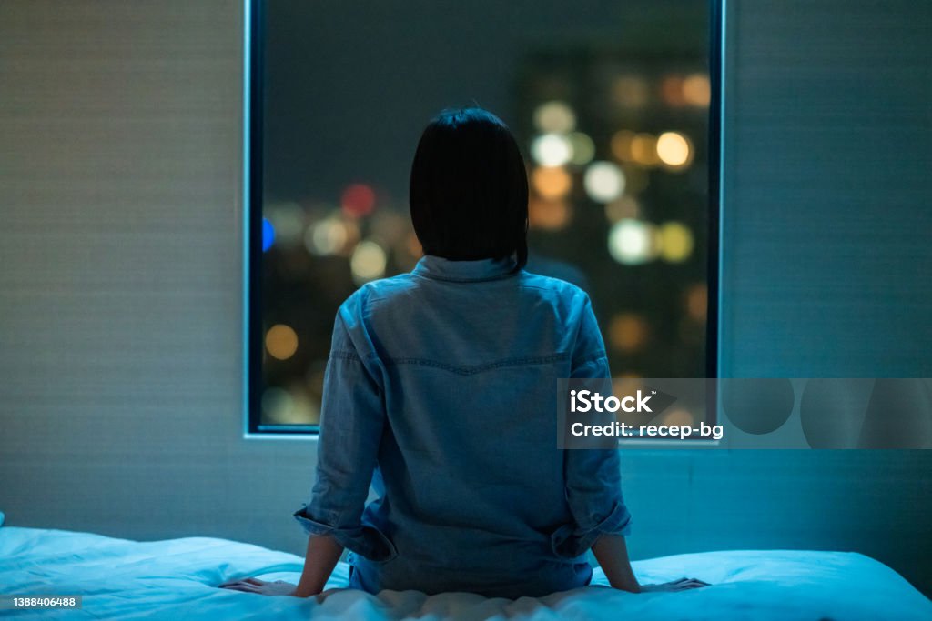 Rear view of woman sitting alone on bed in room and looking through window at night A rear view of a woman sitting alone on a bed in room and looking through the window at night. Insomnia Stock Photo