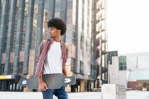 Stock photo of young afro boy walking in the city and carrying his longboard.