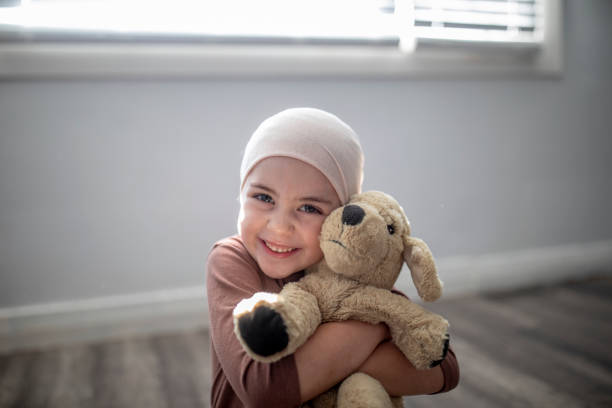 Young Cancer Patient and her Stuffed Animal A sweet little girl battling cancer sits at home on the floor of her bedroom, with her arms wrapped tightly around her stuffed animal as she smiles gently for the portrait. She is dressed casually and wearing a headscarf as the sun shines in on her through the window. girl patient stock pictures, royalty-free photos & images
