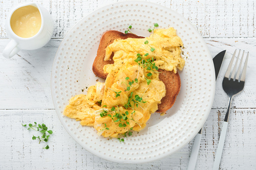 Scrambled Eggs. Fluffy and Buttery scrambled eggs on bread with microgreen radish and hollandaise sauce on white plate over white wooden background. Homemade breakfast or brunch meal. Top view.