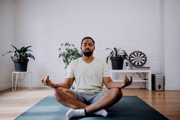 Fitness, meditation and healthy lifestyle concept - black man meditating in lotus pose on exercise mat at home Sporty mid adult black man meditating alone at home, peaceful calm hipster fit guy practicing yoga in lotus pose indoors holding hands in mudra, freedom and calmness concept. meditating stock pictures, royalty-free photos & images