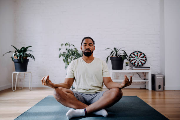 Fitness, meditation and healthy lifestyle concept - black man meditating in lotus pose on exercise mat at home Sporty mid adult black man meditating alone at home, peaceful calm hipster fit guy practicing yoga in lotus pose indoors holding hands in mudra, freedom and calmness concept. mid adult men stock pictures, royalty-free photos & images