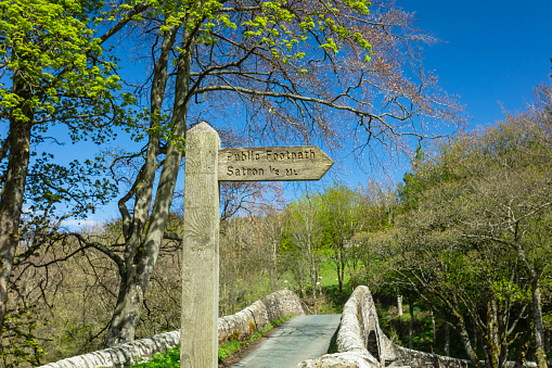 Public footpath sign to Satron, a rural hamlet in Swaledale, with 16th Century Ivelet Bridge in the background.   Springtime in the Yorkshire Dales with blue sky and trees bursting into leaf. Space for copy.