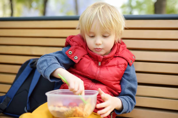 istock Little boy is eating his lunch after kindergarten or school from plastic container on bench in the park. Street take away food for child. Healthy meals for kids. 1388428818