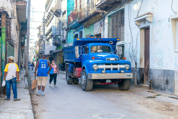 Havana, Cuba - February 12, 2022:  Cuban people with bags walking on a city street where weathered buildings are seen. There are a vintage Ford truck parked in the street.