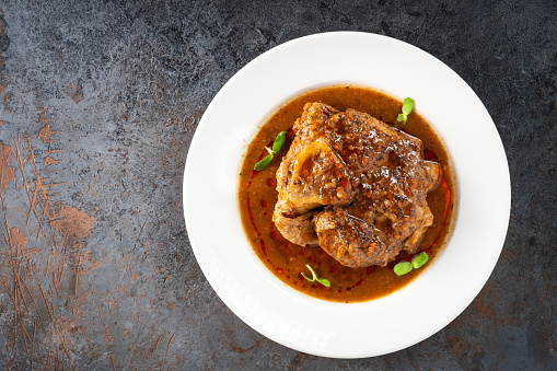 Stewed Osso Buco dish with tomato gravy on a white plate and a stone background, top view