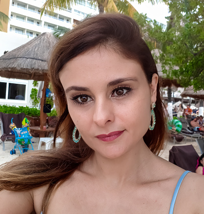 White woman with long brown hair, aquamarine earrings and blue biquini (beachwear) on the beach. Selfie.