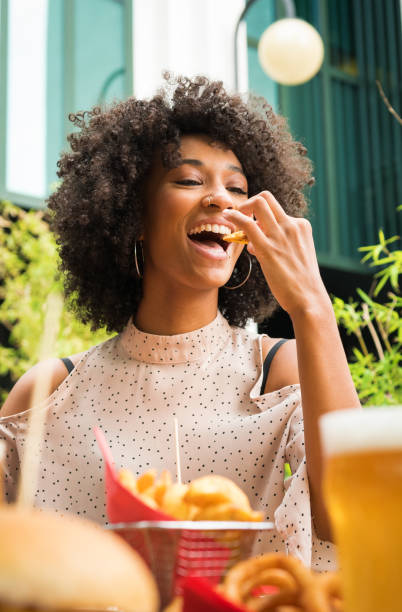 Smiling happy Black woman eating potato wedges in a pub Smiling beautiful young happy Black woman with nose piercing eating potato wedges in a pub in a low angle view over food and glasses of beer snack stock pictures, royalty-free photos & images