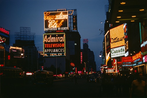 New York City, NY, USA, 1965. The famous Times Square in Manhattan at dusk. Also: pedestrians, neon lights, advertising, shops, buildings and traffic.