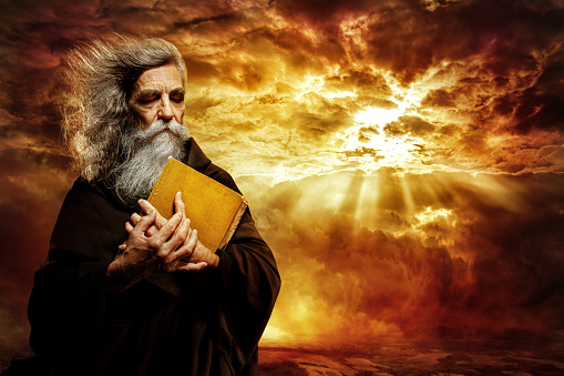 Prophet with Bible. Old Monk with Golden Holy Book praying over Epic Apocalypse Landscape Background. Senior Bearded Man Worship in Black Cloak over Mystery Sunset Sky with Sun Rays shining through Clouds