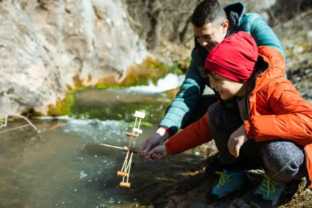 A cute young boy and his dad are holding a water wheel in a mountain stream and learning about sustainable energy